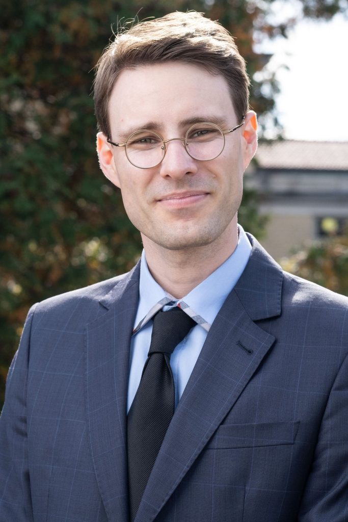 Jean Vilbert wears glasses and a suit, standing in front of a leafy background.