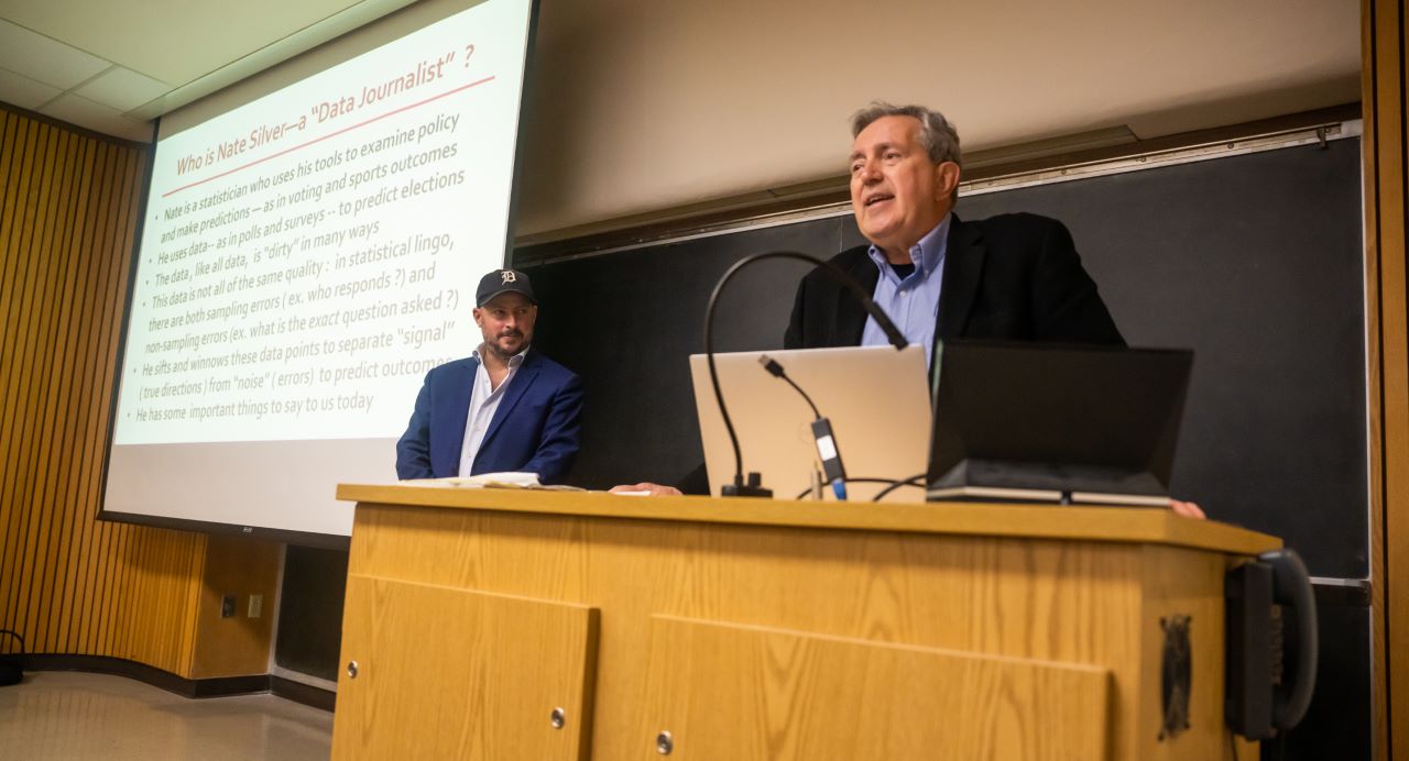 Professor stands at a podium in front of a chalkboard and introduces guest