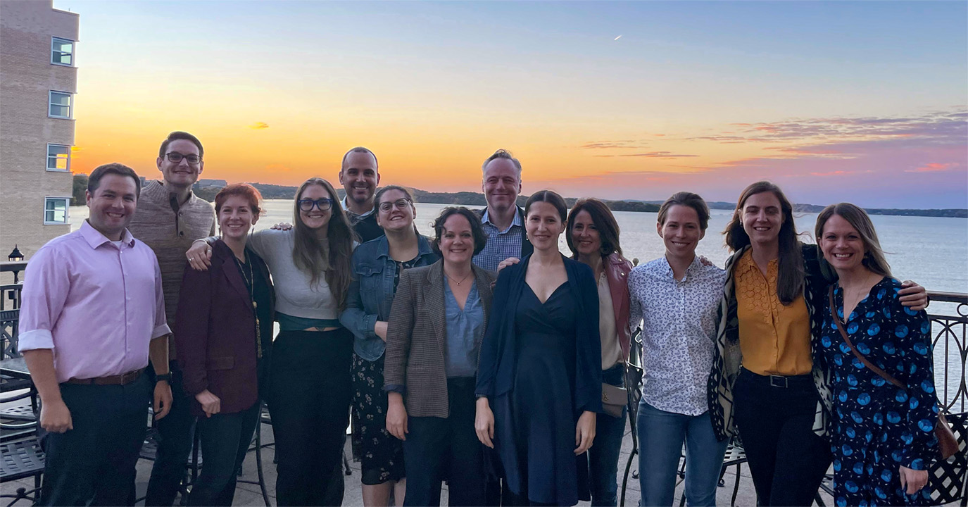 2012 alumni pose for a group photo on a rooftop terrace overlooking a lake at sunset.