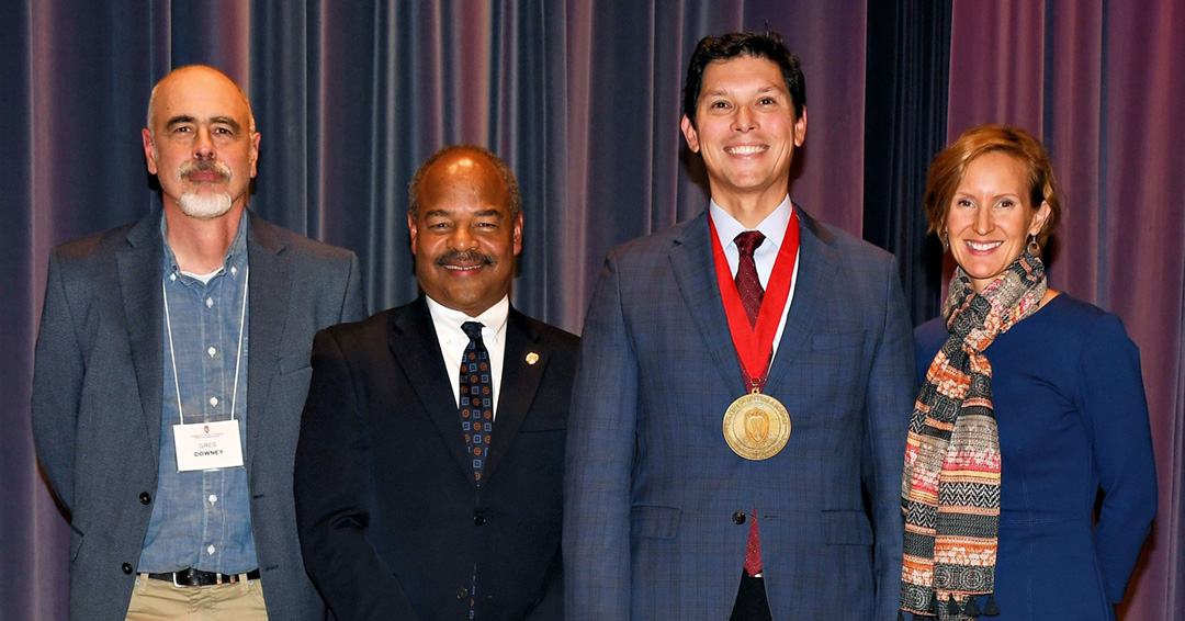 Manny Teodoro wears a medal and stands on a stage with Susan Yackee and College of Letters and Science leadership.