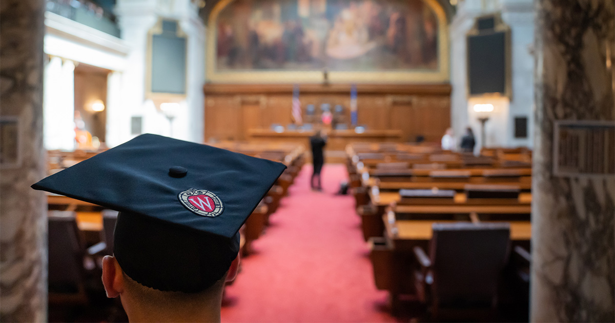 A student wearing a graduation cap waits before a graduation ceremony in the Assembly Chambers of the Wisconsin State Capitol.