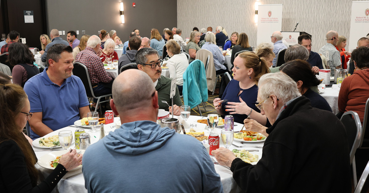People eat dinner and chat at a table in a crowded banquet hall.