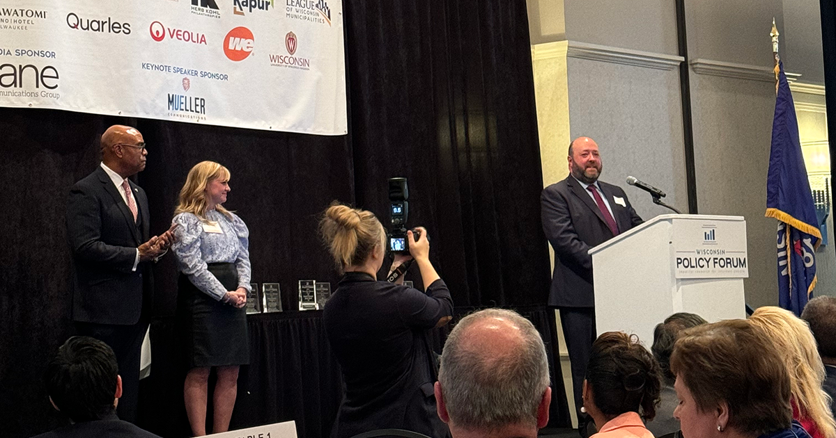 Adam Plotkin speaks at podium while two other people stand on stage