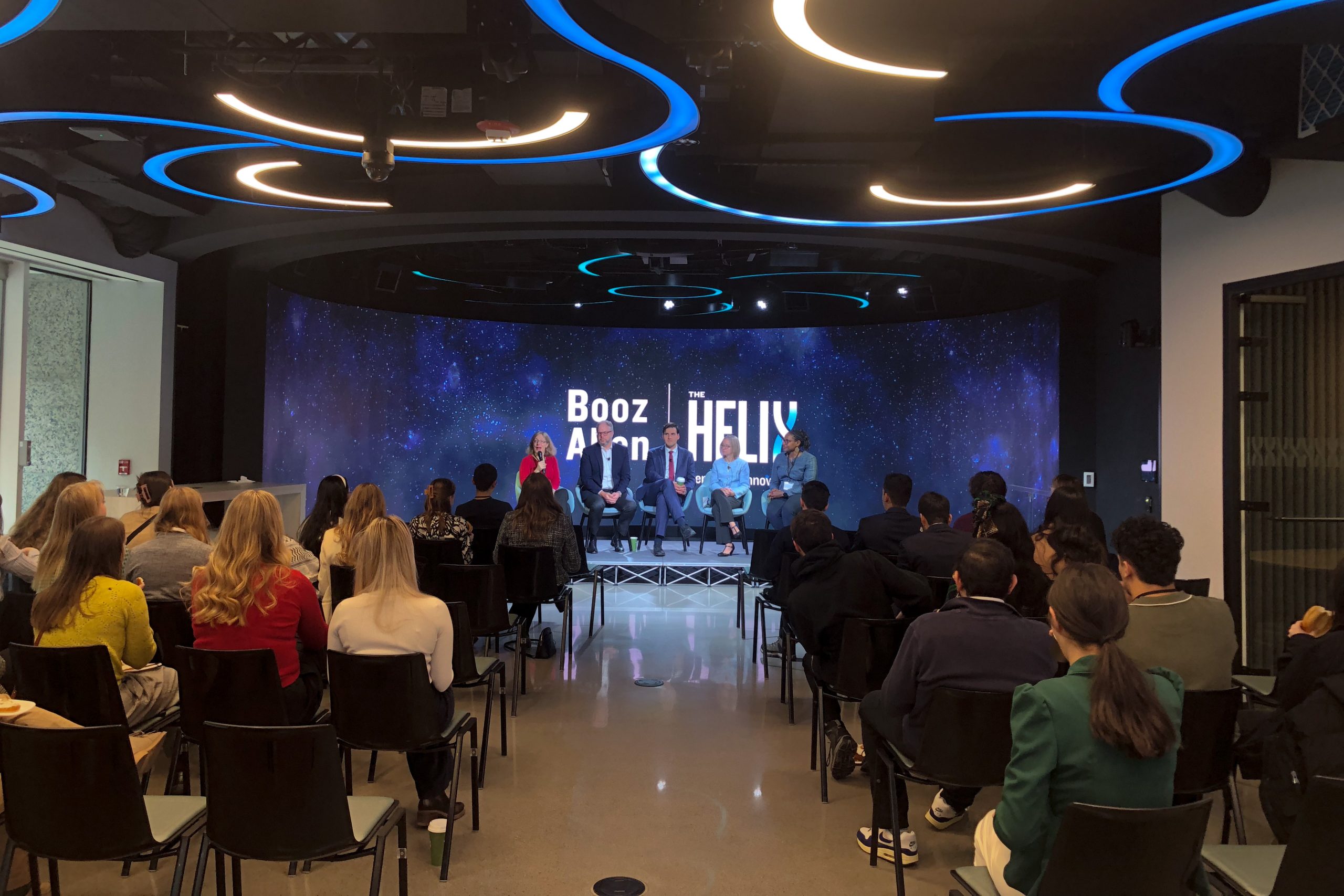 An audience listens to a panel discussion on a stage with the words "Booz Allen" in the background.