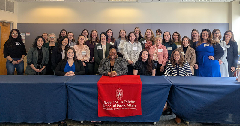 A group of women stand behind a table with a banner that says "Robert M. La Follette School of Public Affairs, University of Wisconsin-Madison"