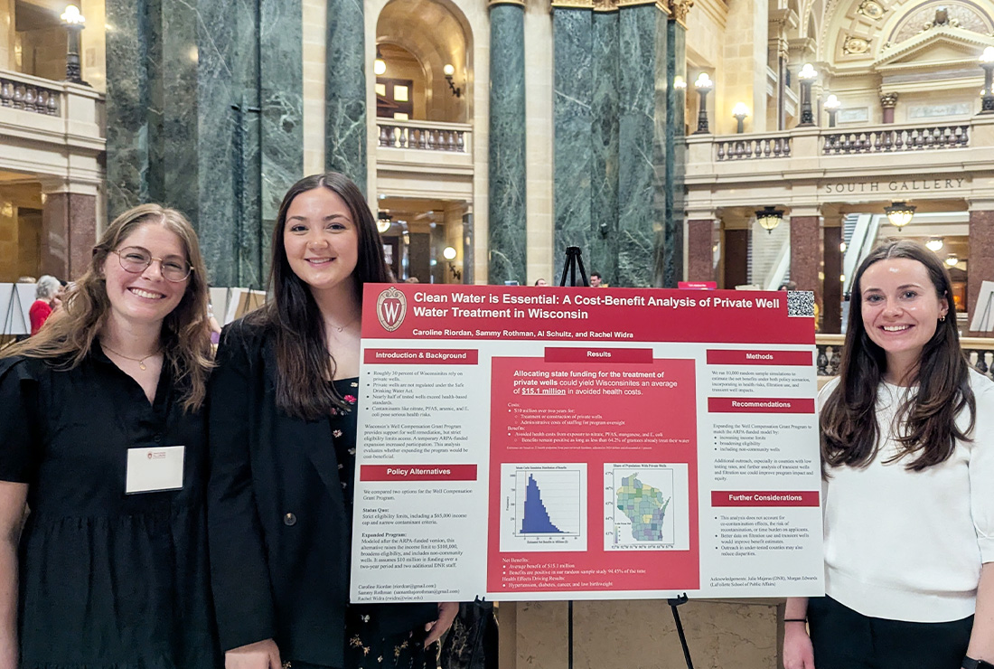 Three graduate students stand in the Wisconsin State Capitol next to a poster they created.