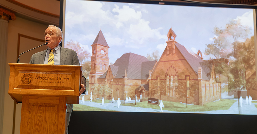 Chuck Pruitt speaks at a podium with a rendering of Herb Kohl Hall visible on a projector screen behind.