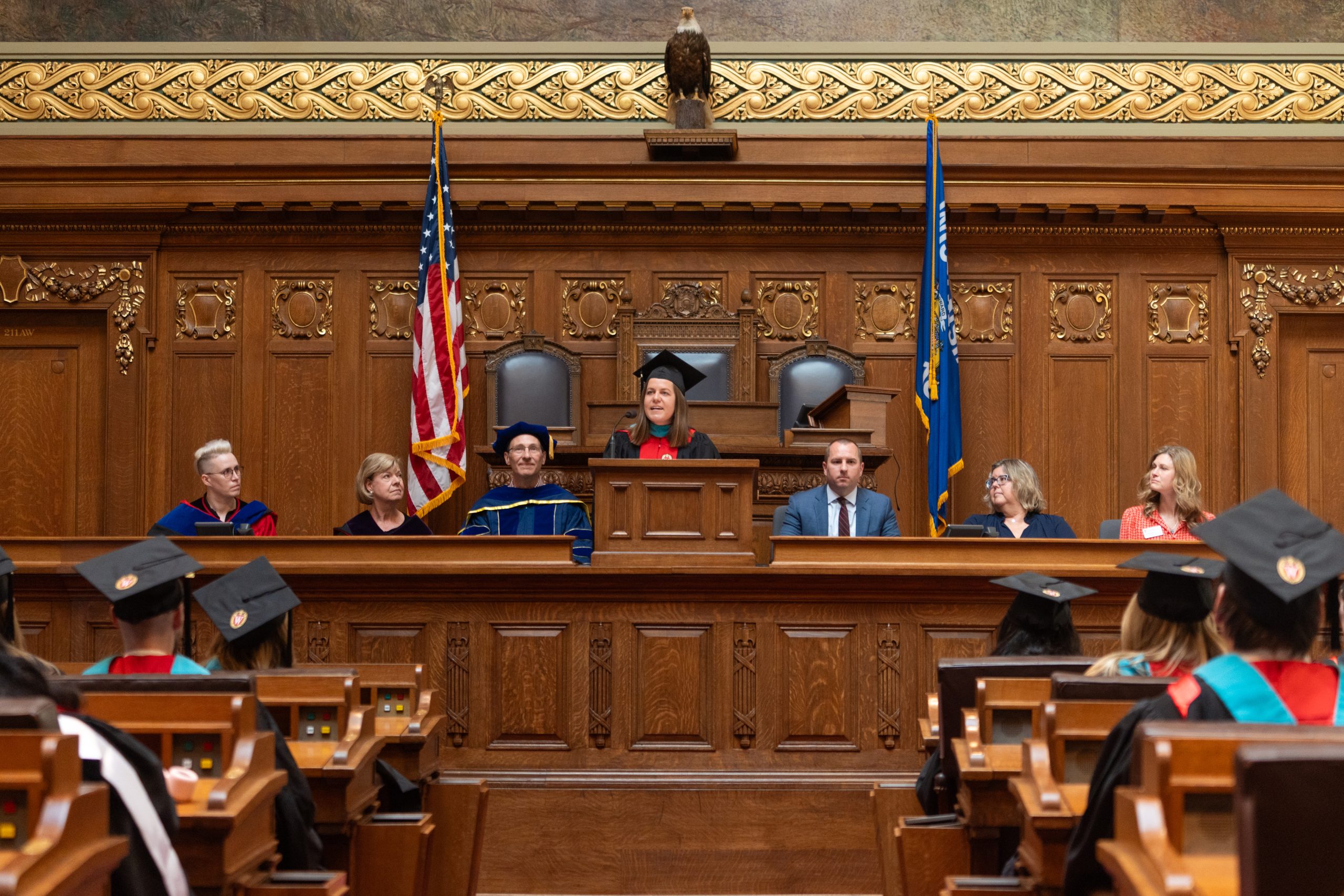 Morgan Pincombe speaks at podium while speakers (left to right) Mariel Barnes, Senator Tammy Baldwin, Jason Delborne, Steve Kulig, Mo O'Connor, and Jennifer Unitan listen with students.