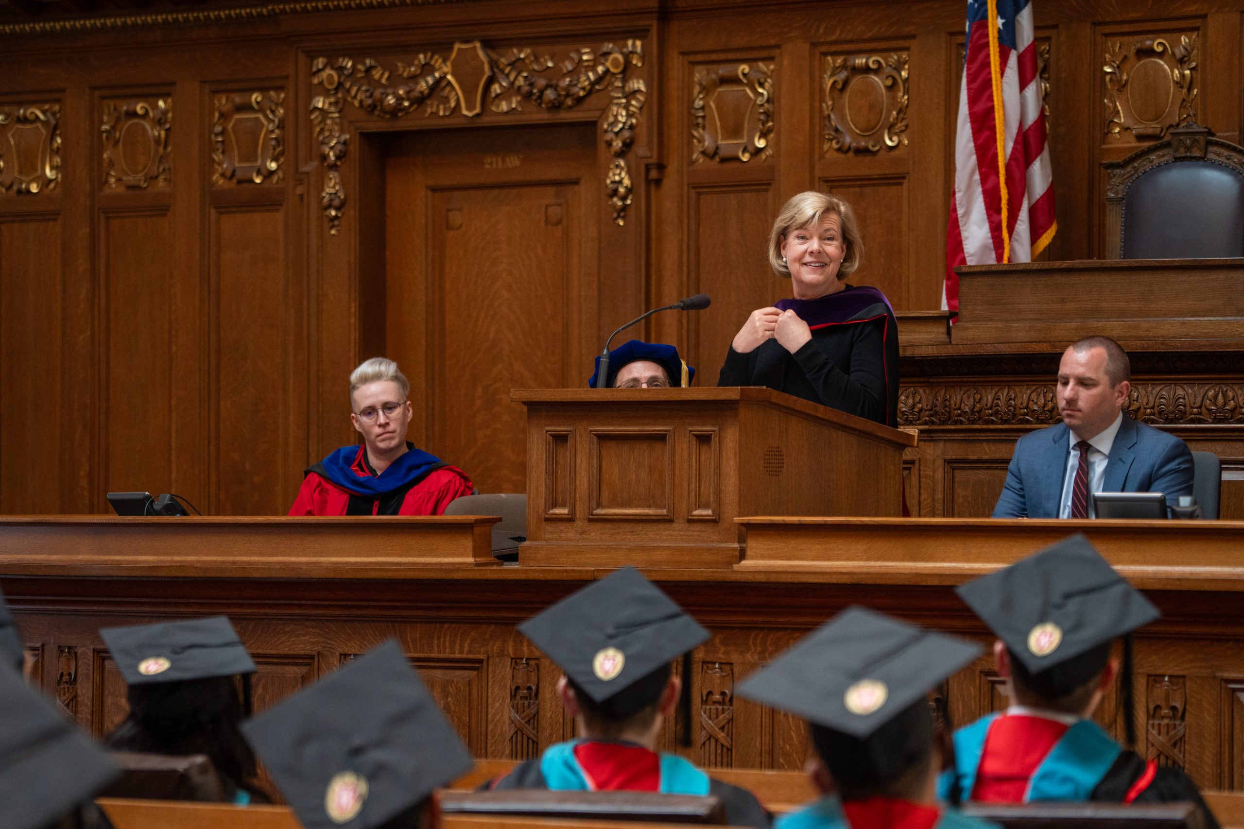Senator Tammy Baldwin speaks at podium as faculty speaker Mariel Barnes, Associate Director Steve Kulig, and students listen