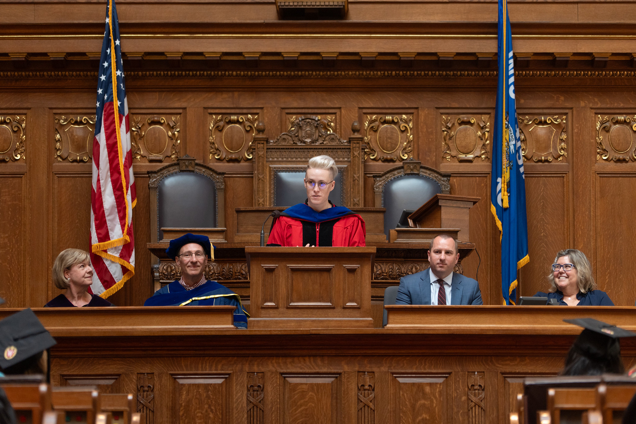 A woman stands at a lectern with two people on each side of her and the American and Wisconsin flags in the background